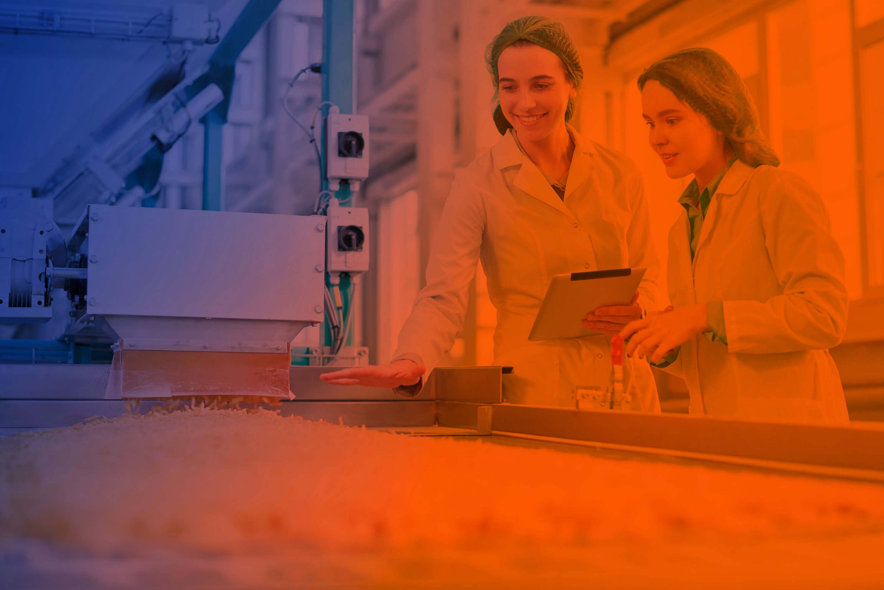 Zwei Mitarbeiterinnen in der Lebensmittelproduktion stehen an der Auslage einer Teigwarenmaschine. Aus einem Trichter fallen frische Teigwaren auf das Band und werden von ihnen geprüft. Sie schauen freundlich. Eine hält einen Tablet-Computer in der Hand.